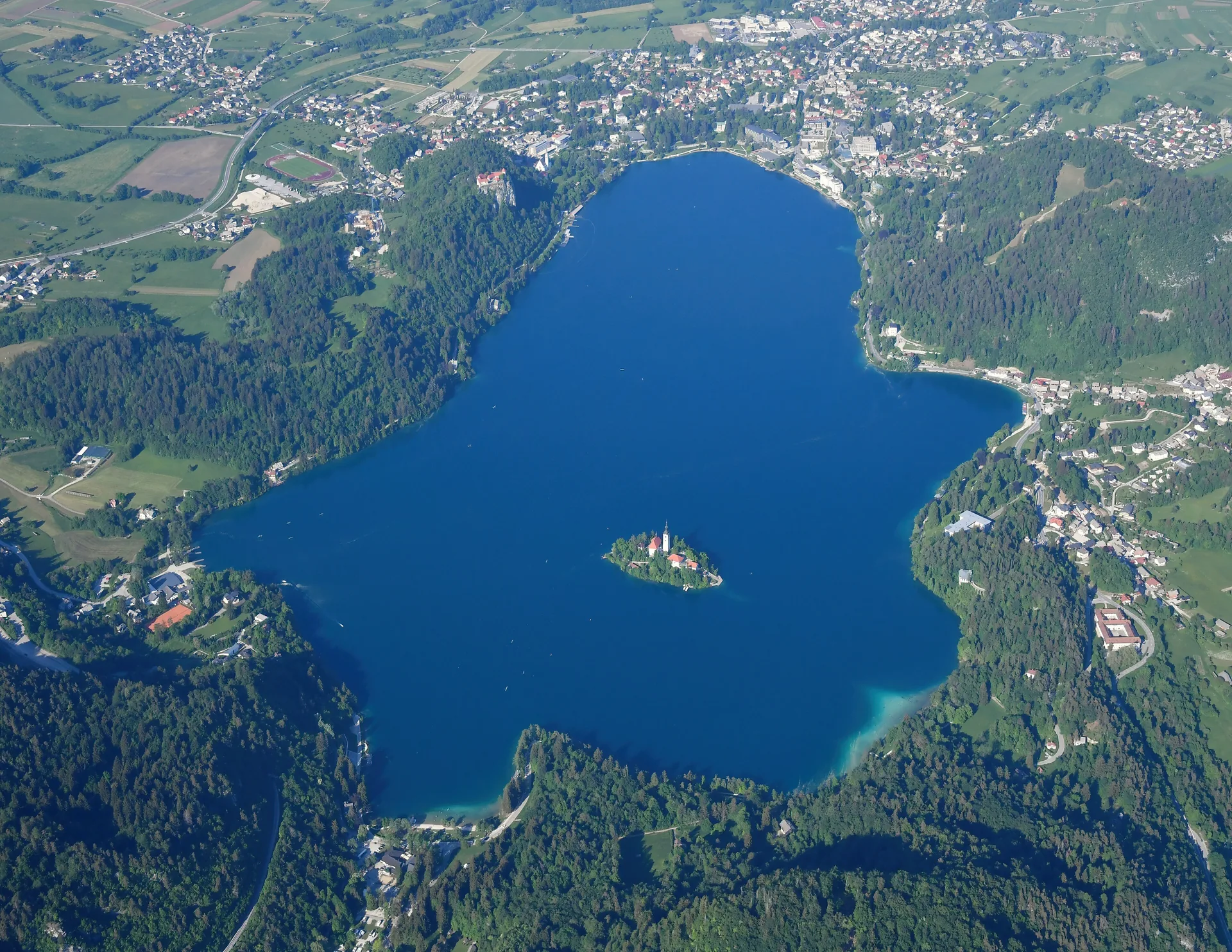 Lake Bled from the air — the island church at the centre of deep blue water, the town and forest around it