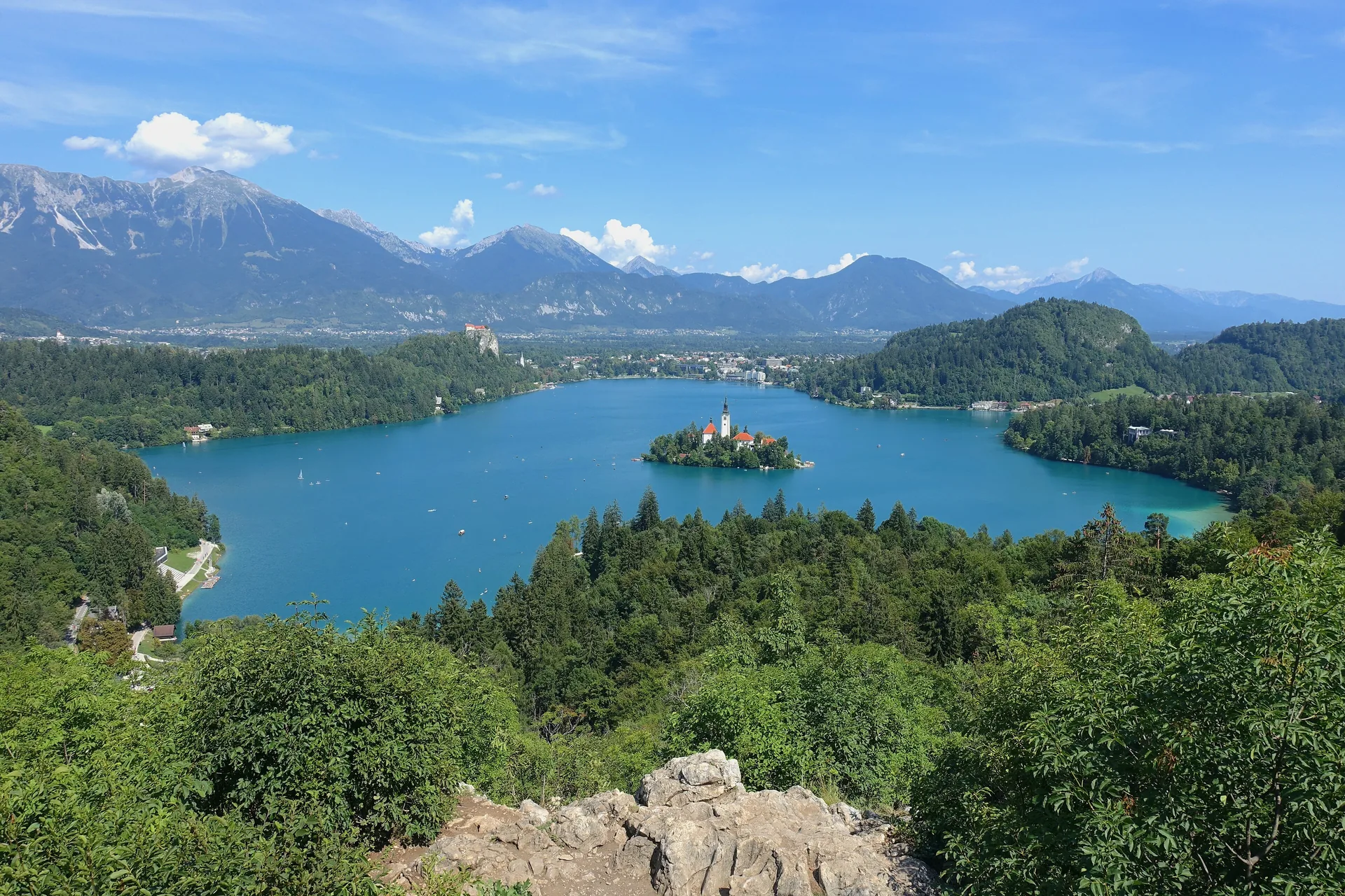 Lake Bled in summer seen from Ojstrica — the island church on the water, the castle on its cliff and the Julian Alps behind
